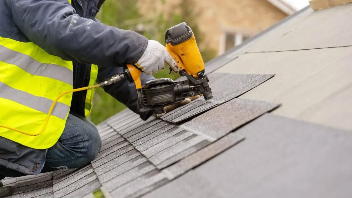 A close-up of a roofer wearing PPE and using a nail gun to install a shingle roof